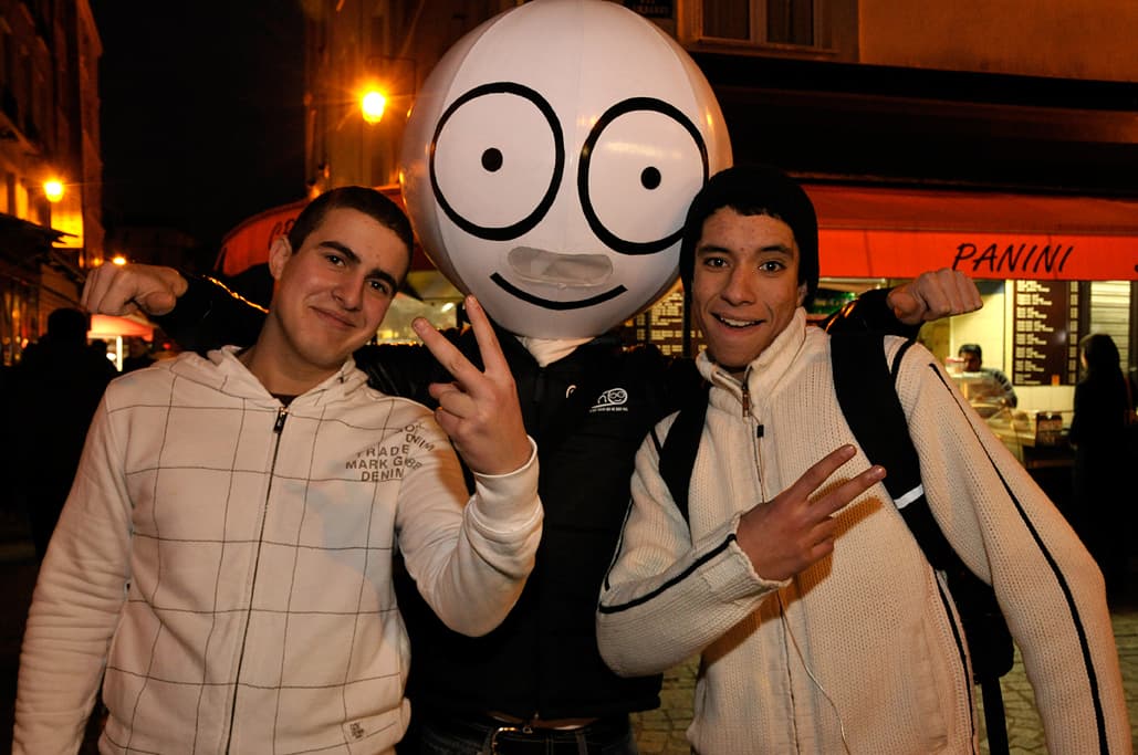 Deux jeunes posant avec une mascotte à tête gonflable souriante, rue parisienne de nuit