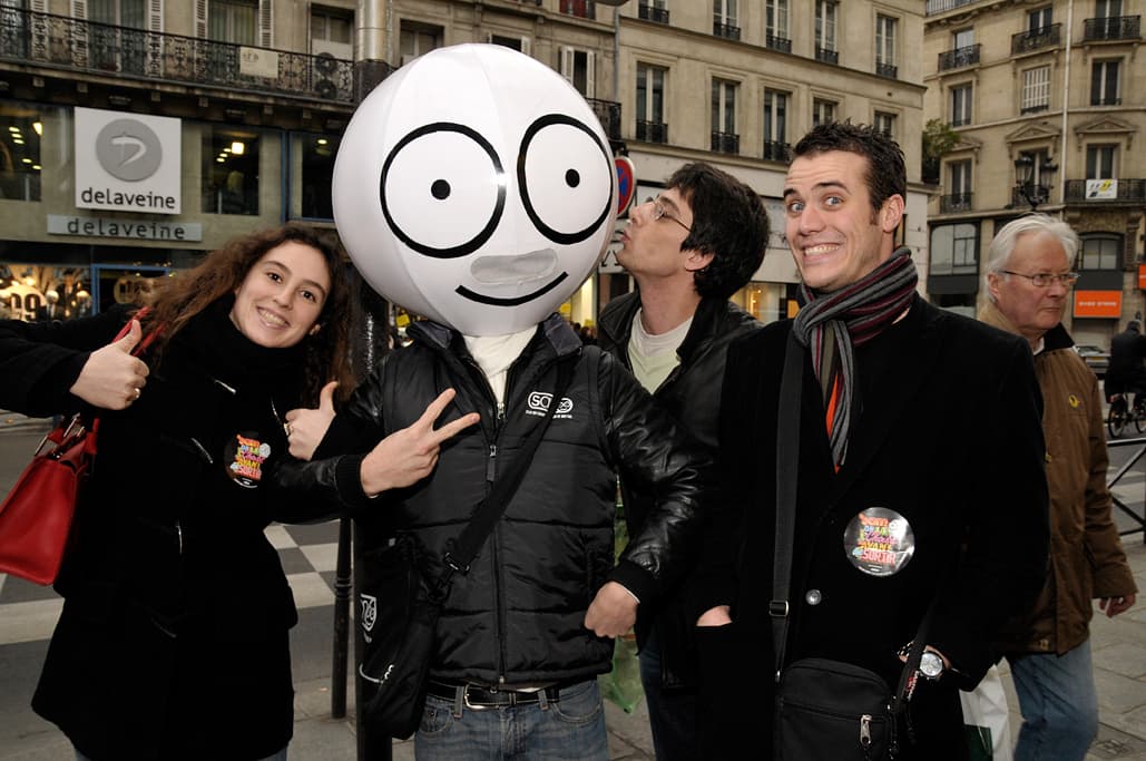 Jeunes souriants posant autour d'une mascotte tête ronde lors d'une opération street marketing dans une rue parisienne