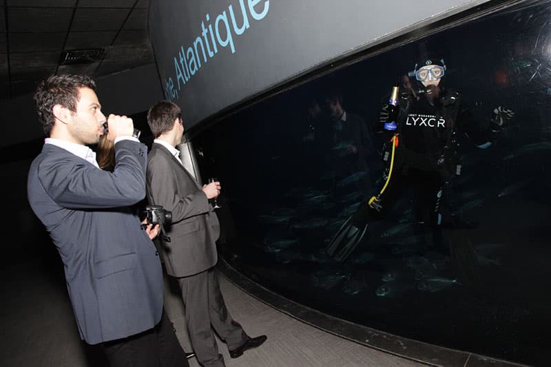 Invités en costume verre à la main devant l'aquarium géant et un plongeur lors de la soirée Lyxor