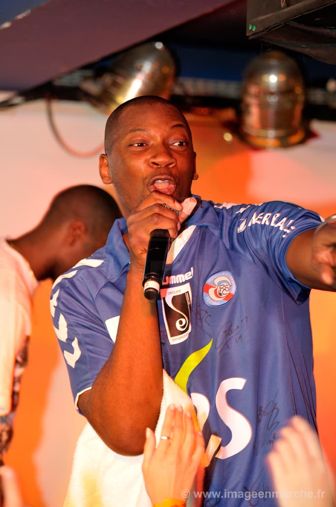 Rappeur en maillot de foot bleu au micro, bras tendu, concert privé