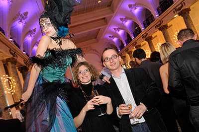Échassière en costume de plumes turquoise et noires posant avec deux invités verre à la main, lors d'une soirée festive d'entreprise dans une salle à colonnes